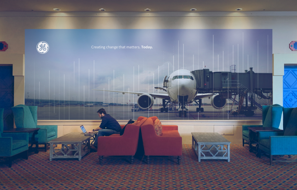 A man is shown sitting on a red couch using his laptop in a large hotel lobby, and behind him along the entire wall is a large poster for GE featuring a passenger jet being loaded with baggage on the tarmac, with the words, "Creating change that matters today". This is an example of environmental branding design; putting your brand in the environment people are existing in. 