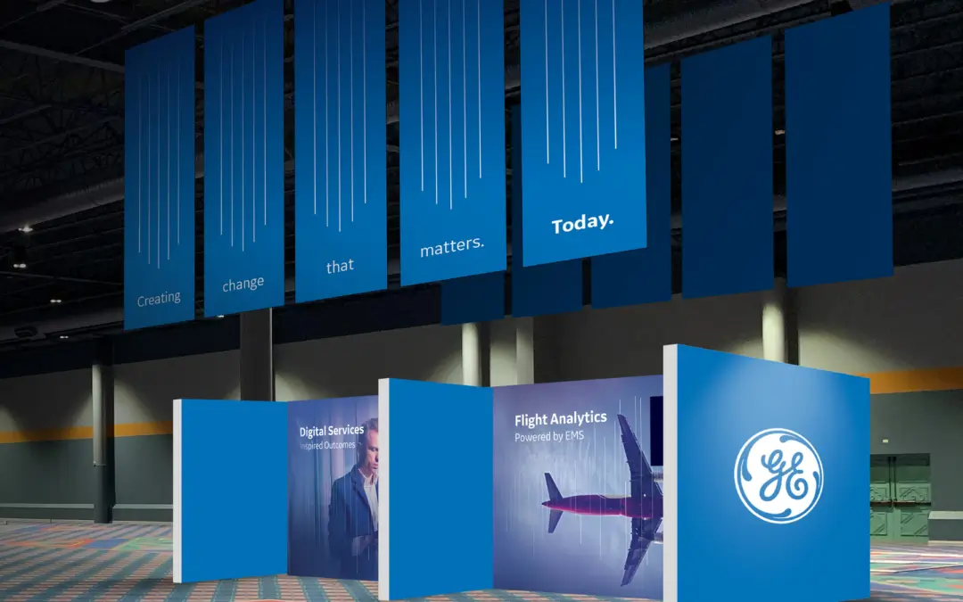 A large lobby area where an environmental branding experience for GE is shown. Posters hanging from the ceiling read "Creating change that matters today" and there are images of people and an airplane on large standing booth graphics on the floor.