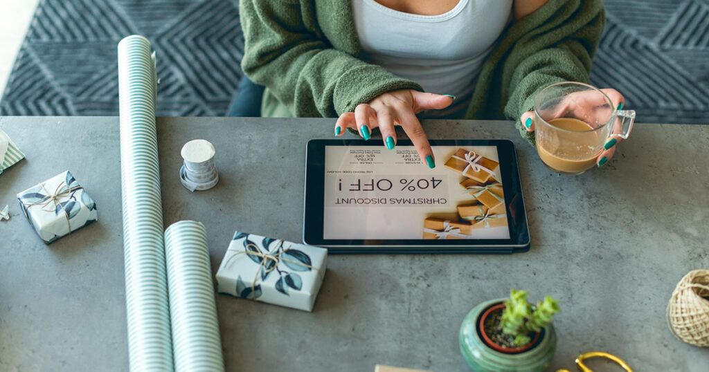 Holiday Shopping Trends 2023 - a woman uses an ipad on a countertop while wrapping christmas presents